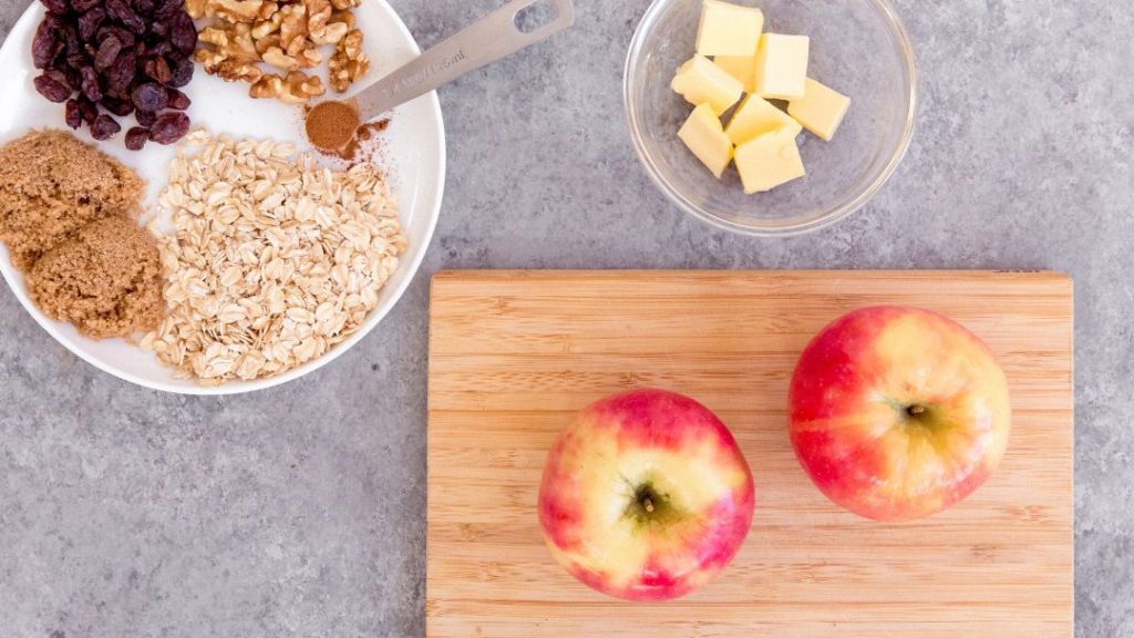 an overhead shot of the ingredients. on a white plate are the dry ingredients, in a clear bowl is butter, and on a chopping board are two apples