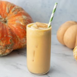 a chai pumpkin spice latte with a green and white striped straw sitting in the glass. There is three pumpkins sitting in the background as decoration.
